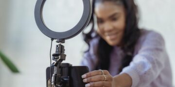 Happy young black woman setting up smartphone before shooting podcast | FintechZoom