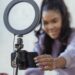 Happy young black woman setting up smartphone before shooting podcast | FintechZoom