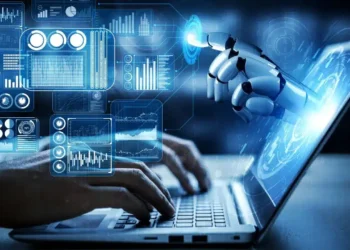A person's hands type on a laptop keyboard. Above the keyboard, a robotic hand reaches towards a holographic interface displaying various graphs, charts, and data visualizations, symbolizing AI integration while highlighting potential AI security threats in technology. | GRAMHIR