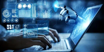 A person's hands type on a laptop keyboard. Above the keyboard, a robotic hand reaches towards a holographic interface displaying various graphs, charts, and data visualizations, symbolizing AI integration while highlighting potential AI security threats in technology. | GRAMHIR