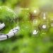 A robotic hand holds a transparent globe, with various icons and data points radiating from it, including GPS, temperature, PH scale, and agricultural symbols. The background features a blurred green nature setting, seamlessly blending Climate AI technology with the environment. | GRAMHIR