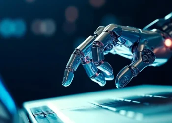 A robotic hand with intricate metallic details reaches out to touch the keyboard of a laptop, emitting a soft glow in a dimly lit environment. The scene evokes a sense of futuristic technology and artificial intelligence, reminiscent of the advancements driving AI stocks today. | GRAMHIR