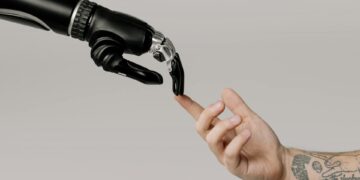 A robotic hand, embodying generalist intelligence, reaches out to gently touch the tattooed finger of a human. The neutral background highlights the profound connection between advanced technology and humanity. | GRAMHIR