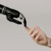 A robotic hand, embodying generalist intelligence, reaches out to gently touch the tattooed finger of a human. The neutral background highlights the profound connection between advanced technology and humanity. | GRAMHIR
