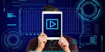 A person holds a tablet in front of their face, displaying a play button. The background is adorned with digital interface elements—binary code, radial graphs, and futuristic tech designs in blue tones—alluding to tools that help make high-quality videos. | GRAMHIR