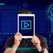 A person holds a tablet in front of their face, displaying a play button. The background is adorned with digital interface elements—binary code, radial graphs, and futuristic tech designs in blue tones—alluding to tools that help make high-quality videos. | GRAMHIR