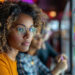 A person with glasses and curly hair is intently looking at a board filled with colorful charts and graphs. They are wearing a yellow shirt. Another person in the background is pointing at the board. The setting appears to be a modern office or study area. | GRAMHIR