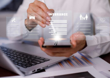 A person uses a stylus on a smartphone, interacting with AI in modern insurance software. Holographic icons—wrench, car, family, heart—float above, symbolizing various insurances. A laptop and paperwork sit in the background, illustrating the seamless integration of technology. | GRAMHIR