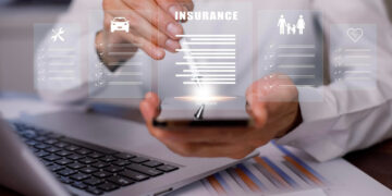 A person uses a stylus on a smartphone, interacting with AI in modern insurance software. Holographic icons—wrench, car, family, heart—float above, symbolizing various insurances. A laptop and paperwork sit in the background, illustrating the seamless integration of technology. | GRAMHIR