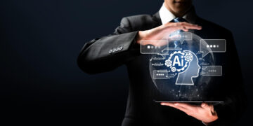 A man in a suit, embodying an AI consultant, holds a digital tablet displaying a glowing AI hologram shaped like a human profile. The hologram features circuit patterns and icons against a dark background. | GRAMHIR