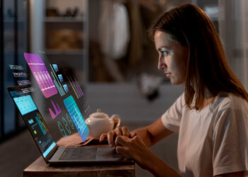 A woman sits at a desk using a laptop with colorful digital data graphs and charts projected in the air above the keyboard, suggesting advanced technology or augmented reality. A teapot and cup are also on the table. | GRAMHIR