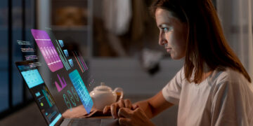 A woman sits at a desk using a laptop with colorful digital data graphs and charts projected in the air above the keyboard, suggesting advanced technology or augmented reality. A teapot and cup are also on the table. | GRAMHIR