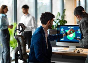 Two people review data from Employee Monitoring Software on a computer screen at a desk, while two others chat in the background of a modern, bright office space filled with large windows and plants. | GRAMHIR