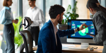 Two people review data from Employee Monitoring Software on a computer screen at a desk, while two others chat in the background of a modern, bright office space filled with large windows and plants. | GRAMHIR