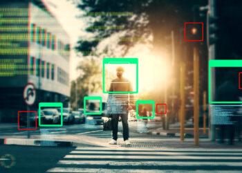 A person crossing a city street at a crosswalk is highlighted by digital green and red rectangles, symbolizing geospatial annotation and object recognition technology analyzing the scene. | GRAMHIR