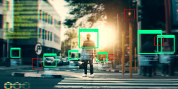 A person crossing a city street at a crosswalk is highlighted by digital green and red rectangles, symbolizing geospatial annotation and object recognition technology analyzing the scene. | GRAMHIR