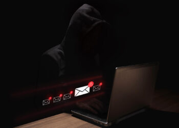 A person in a dark hoodie sits at a laptop in a dimly lit setting, with illuminated email icons and warning symbols appearing in front of the screen, suggesting a cybersecurity or phishing threat. | GRAMHIR