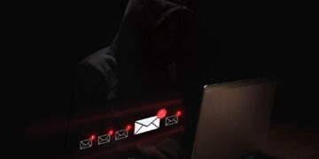 A person in a dark hoodie sits at a laptop in a dimly lit setting, with illuminated email icons and warning symbols appearing in front of the screen, suggesting a cybersecurity or phishing threat. | GRAMHIR