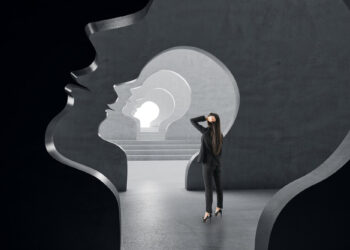 A person stands inside a series of large, nested human head silhouettes, carved into a dark stone wall. The silhouetted heads create a tunnel effect leading to CerebrumIQ's bright light in the distance. The person looks towards it, holding their head in contemplation. | GRAMHIR