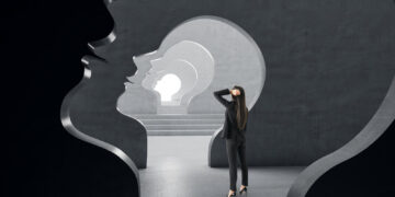 A person stands inside a series of large, nested human head silhouettes, carved into a dark stone wall. The silhouetted heads create a tunnel effect leading to CerebrumIQ's bright light in the distance. The person looks towards it, holding their head in contemplation. | GRAMHIR