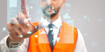 A construction professional in an orange vest and hard hat interacts with a virtual screen displaying digital icons related to construction, such as buildings, tools, and graphs. The focus is on the digital interface powered by an EHS Management Tool with Artificial Intelligence. | GRAMHIR