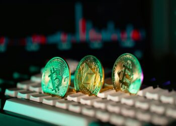 Three cryptocurrency coins—Bitcoin, Ethereum, and Litecoin—stand upright on a computer keyboard, with a blurred digital graph in the background. The scene’s vivid lighting hints at debates like “Will AI Replace Crypto?” in the digital finance world. | GRAMHIR