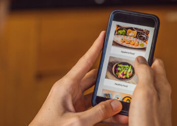 A person holding a smartphone and browsing a food delivery app, which showcases sushi and healthy food dishes on the screen—an example of how mobile optimization tactics enhance user experience. | GRAMHIR