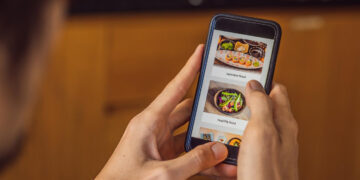 A person holding a smartphone and browsing a food delivery app, which showcases sushi and healthy food dishes on the screen—an example of how mobile optimization tactics enhance user experience. | GRAMHIR