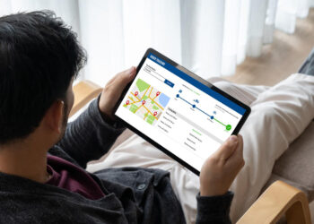 A person sitting on a chair holding a tablet that displays a delivery tracking map and progress timeline, showing the package’s location and estimated delivery steps. | GRAMHIR