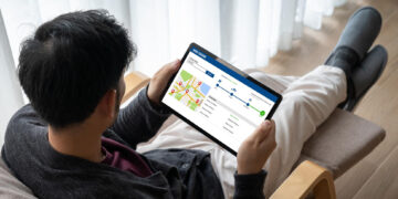 A person sitting on a chair holding a tablet that displays a delivery tracking map and progress timeline, showing the package’s location and estimated delivery steps. | GRAMHIR