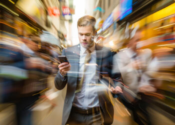 A man in a suit looks at his phone while walking through a busy city street, the blurred background emphasizing the fast-paced environment—an ironic contrast to someone searching for tips on how to plan a stress-free digital detox. | GRAMHIR