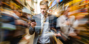 A man in a suit looks at his phone while walking through a busy city street, the blurred background emphasizing the fast-paced environment—an ironic contrast to someone searching for tips on how to plan a stress-free digital detox. | GRAMHIR