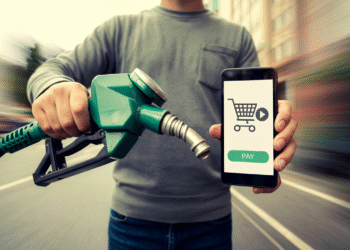 A person holds a fuel pump nozzle in one hand and a smartphone in the other, displaying a shopping cart icon, "pay" button, and Gas Card option, suggesting mobile payment for fuel. The background is blurred. | GRAMHIR