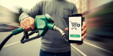 A person holds a fuel pump nozzle in one hand and a smartphone in the other, displaying a shopping cart icon, "pay" button, and Gas Card option, suggesting mobile payment for fuel. The background is blurred. | GRAMHIR