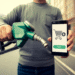A person holds a fuel pump nozzle in one hand and a smartphone in the other, displaying a shopping cart icon, "pay" button, and Gas Card option, suggesting mobile payment for fuel. The background is blurred. | GRAMHIR