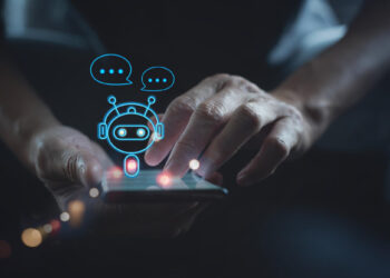 A person uses a smartphone with a digital illustration of a chatbot, depicted as a small robot with speech bubbles hovering above the screen, representing AI Chatbots and advanced communication technology. | GRAMHIR