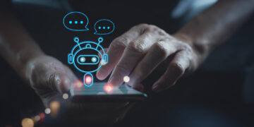 A person uses a smartphone with a digital illustration of a chatbot, depicted as a small robot with speech bubbles hovering above the screen, representing AI Chatbots and advanced communication technology. | GRAMHIR