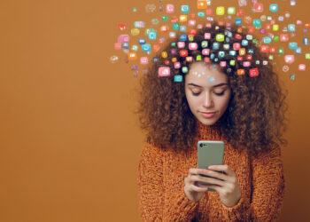 A young woman with curly hair in an orange sweater looks at her smartphone, colorful app icons floating above her head on a solid brown background, suggesting digital thoughts or introspective tools designed for individuals seeking emotional orientation. | GRAMHIR