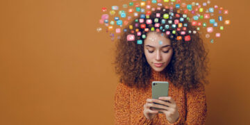 A young woman with curly hair in an orange sweater looks at her smartphone, colorful app icons floating above her head on a solid brown background, suggesting digital thoughts or introspective tools designed for individuals seeking emotional orientation. | GRAMHIR