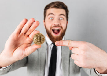 A bearded man in a suit excitedly points to a gold Bitcoin coin he's holding up, with a wide smile and surprised expression against a plain gray background. | GRAMHIR