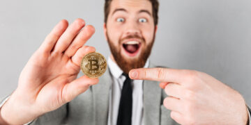 A bearded man in a suit excitedly points to a gold Bitcoin coin he's holding up, with a wide smile and surprised expression against a plain gray background. | GRAMHIR