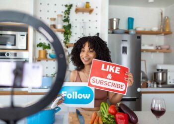 A woman in a kitchen holds “LIKE & SUBSCRIBE” and “Follow” signs while recording a video with a smartphone on a ring light. Vegetables, utensils, and a glass of red wine are on the counter in front of her. | GRAMHIR