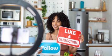 A woman in a kitchen holds “LIKE & SUBSCRIBE” and “Follow” signs while recording a video with a smartphone on a ring light. Vegetables, utensils, and a glass of red wine are on the counter in front of her. | GRAMHIR