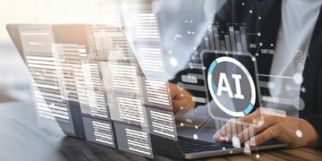 A person uses a laptop at a desk, with digital overlays of data, charts, and a large AI symbol on the screen, representing artificial intelligence technology. | GRAMHIR