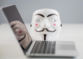 A Guy Fawkes mask is placed behind an open laptop on a white table, with the mask’s reflection visible on the laptop screen, symbolizing anonymity and hacking. | GRAMHIR