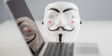 A Guy Fawkes mask is placed behind an open laptop on a white table, with the mask’s reflection visible on the laptop screen, symbolizing anonymity and hacking. | GRAMHIR