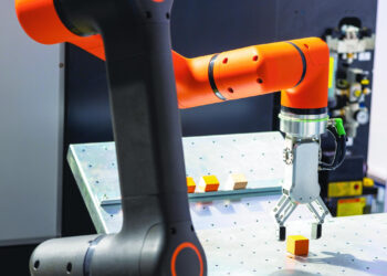 An orange and black robotic arm with a gripper is picking up a small yellow cube from a perforated metal surface, with other cubes on the table in a technological lab setting. | GRAMHIR