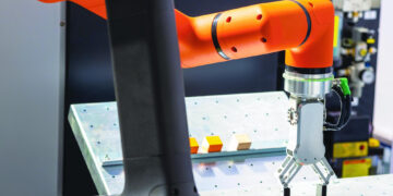 An orange and black robotic arm with a gripper is picking up a small yellow cube from a perforated metal surface, with other cubes on the table in a technological lab setting. | GRAMHIR