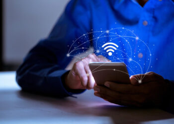 A person in a blue shirt uses a smartphone, with a glowing Wi-Fi symbol and network lines digitally illustrated above the device, suggesting wireless connectivity or internet access. | GRAMHIR