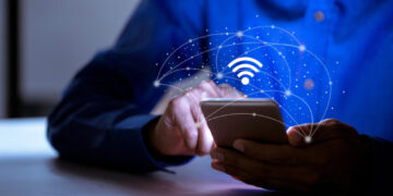 A person in a blue shirt uses a smartphone, with a glowing Wi-Fi symbol and network lines digitally illustrated above the device, suggesting wireless connectivity or internet access. | GRAMHIR